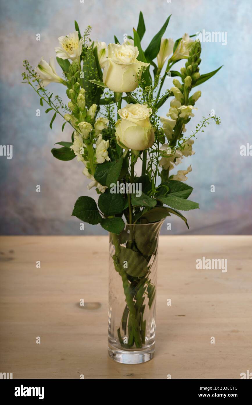 Weiße Rosen und verschiedene weiße Blumen in einer Anordnung in einer Glasvase auf einem Kieferntisch. Stockfoto