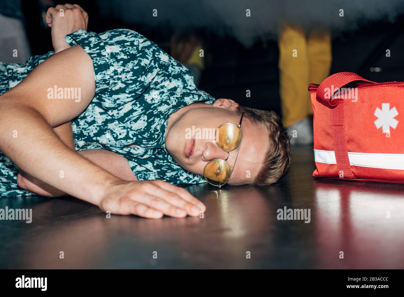 Kranker Mann in Sonnenbrille auf dem Boden in der Nähe der Erste-Hilfe-Kit-Tasche im Nachtclub Stockfoto