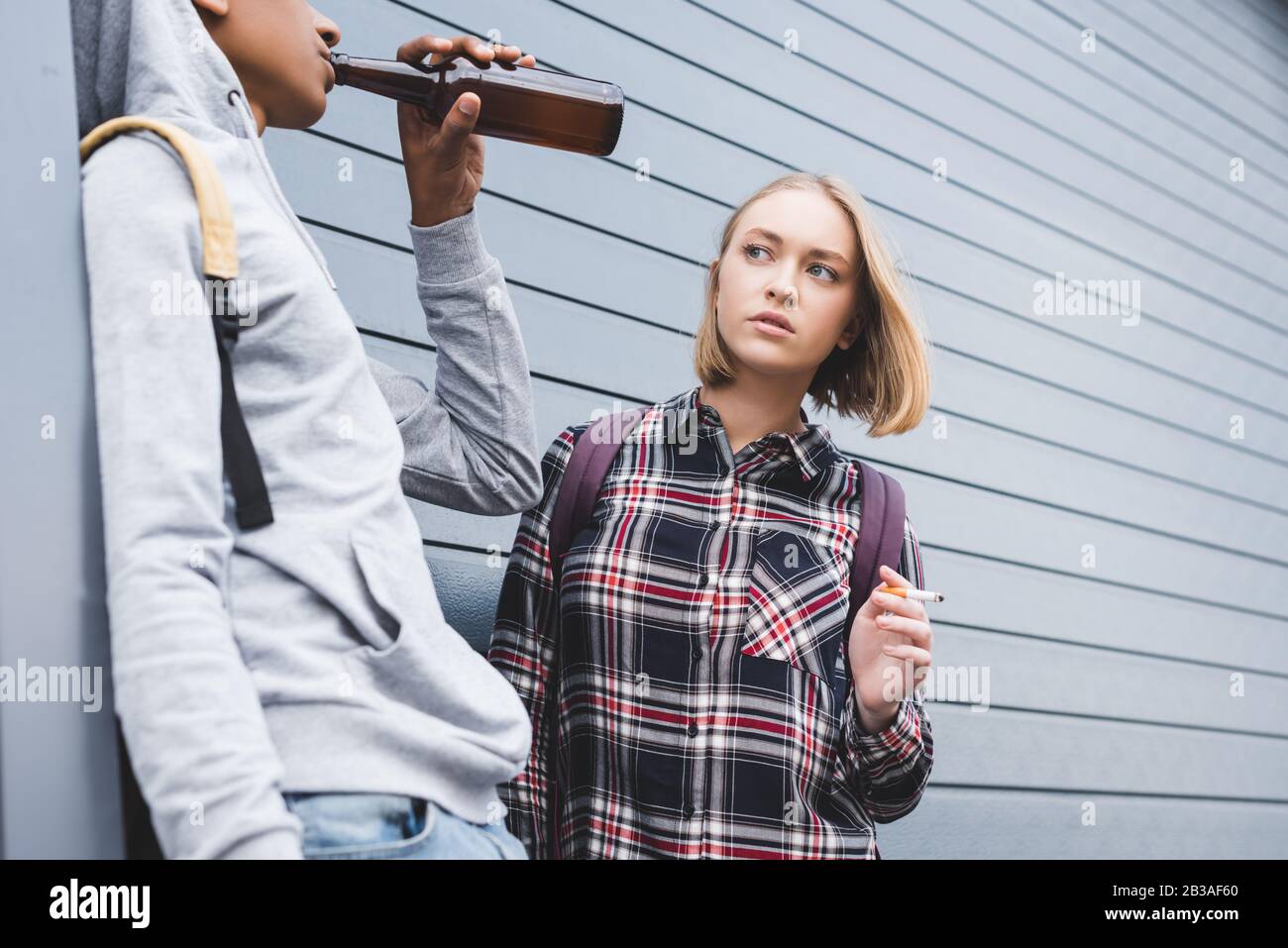 afroamerikanischer Junge und blonde Teenager, die wegschauen, Zigarette rauchen und Bier halten Stockfoto