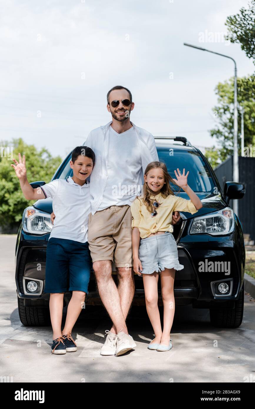 Glücklicher Vater in einer Sonnenbrille, die mit Kindern steht und die Hände in der Nähe des Autos winkt Stockfoto