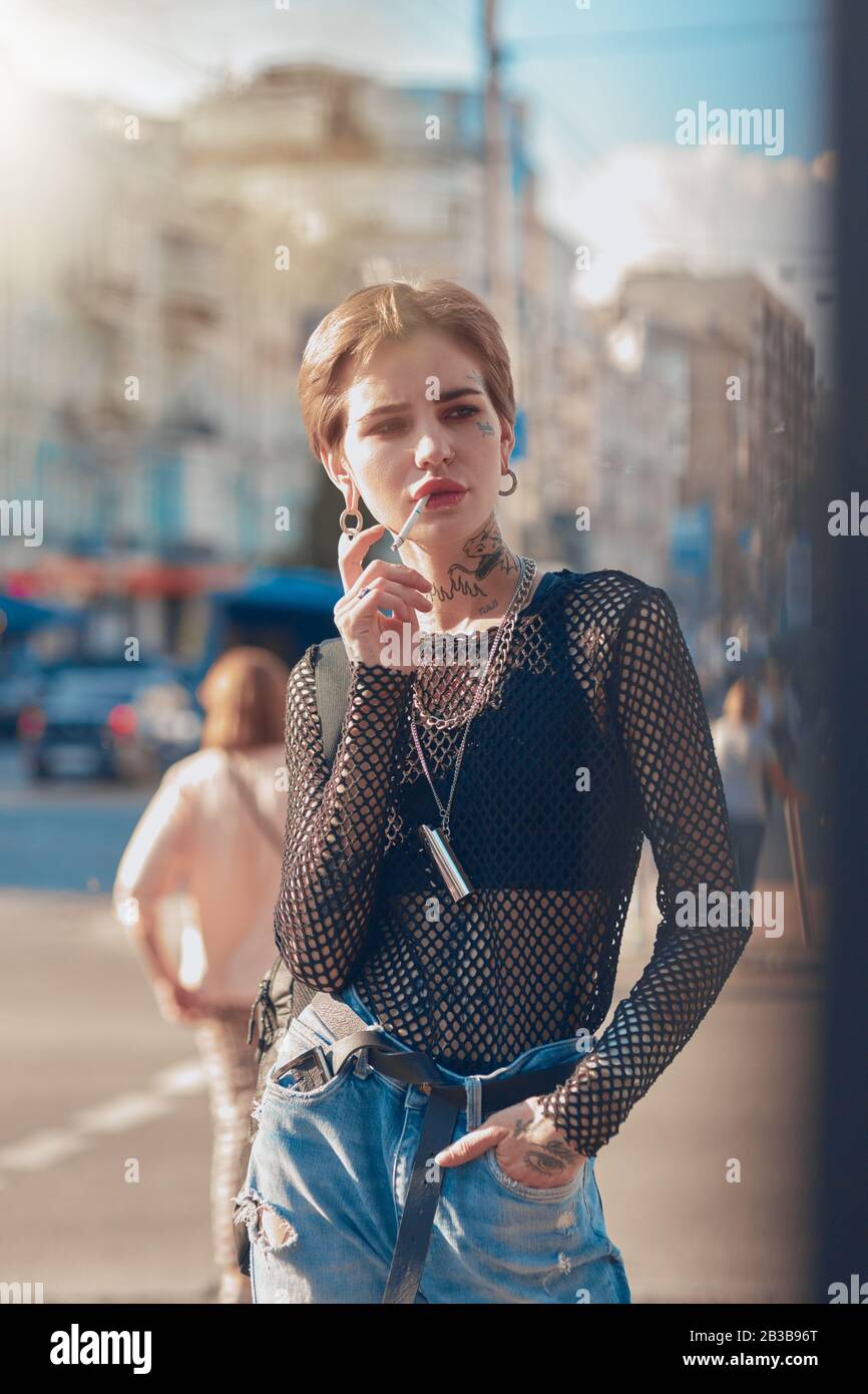Urbaner Stil. Junge, stilvolle Frau, die im Freien Hand in der Tasche steht, raucht eine Zigarette, die aufdringlich aussieht Stockfoto