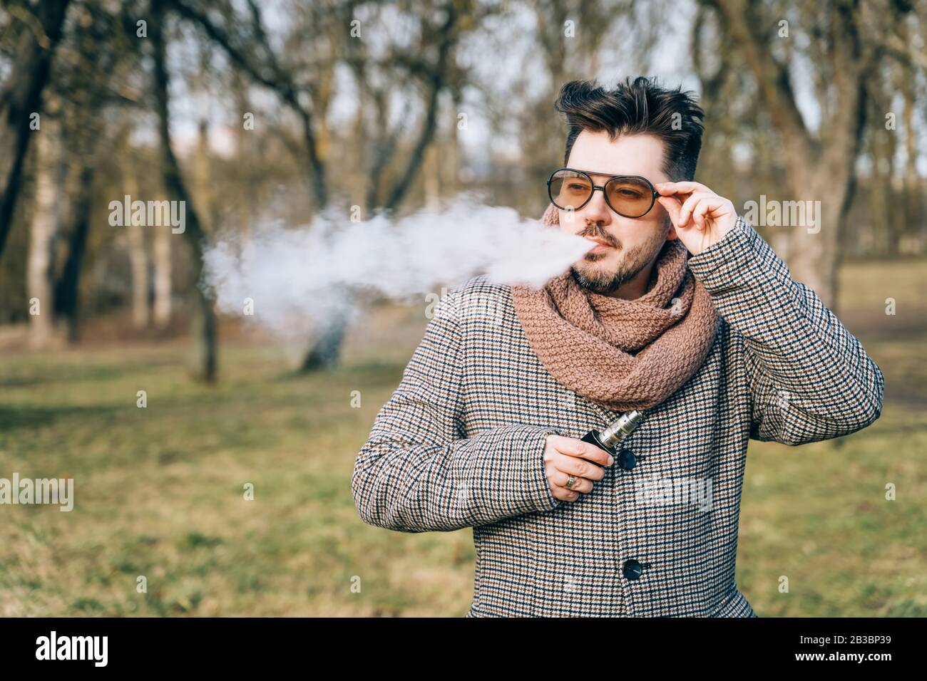 Selbstbewusster junger bärtiger Mann in Sonnenbrille schäumt elektronische Zigarette. Ein junger, hübsch bärtiger Flusspferd, der im Park eine E-Zigarette im Freien umgibt Stockfoto