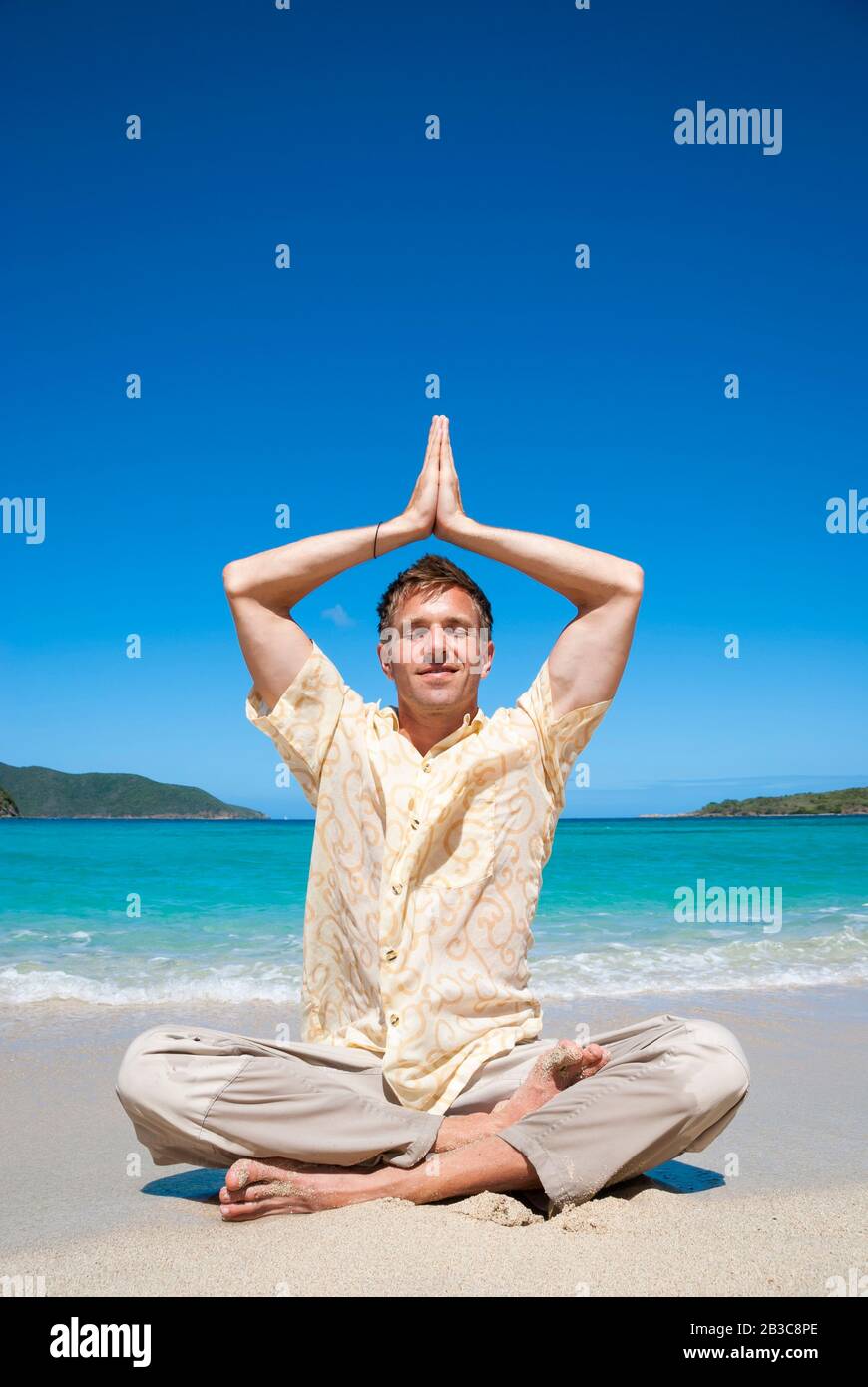 Lächelnder Mann, der in leichter Yoga-Pose sitzt und namaste Hände über dem Kopf an der Kronenkakra an einem tropischen Strand hat Stockfoto