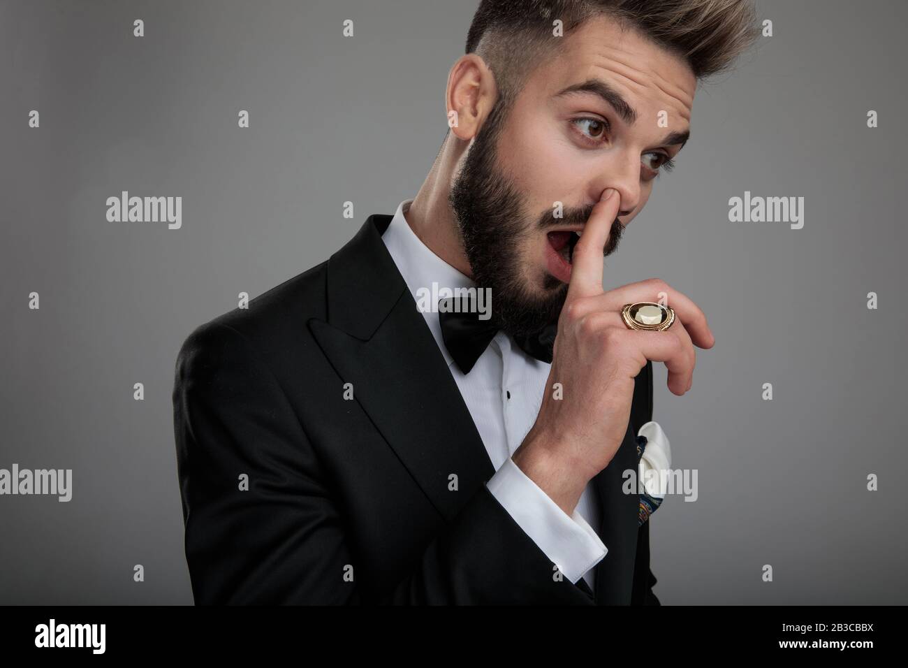 Nahaufnahme eines tollpatschigen Bräutigam, der seine Nase pflückt und seinen Ring zeigt, während er Tuxedo und Bowtie trägt und auf grauem Studiohintergrund steht Stockfoto