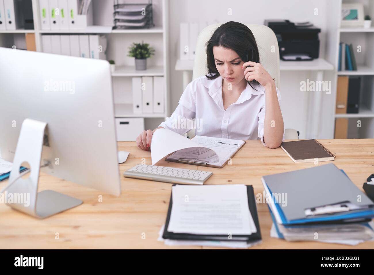 Konzentrierte junge Frau in weißer Bluse, die am Holztisch sitzt und die Papiere in der Zwischenablage analysiert Stockfoto