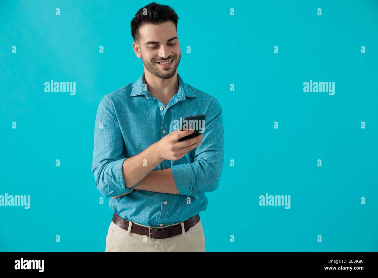 Der hübsche, legere Mann in blauem Hemd steht mit gekreuzten Armen, während er auf dem blauen Studiohintergrund auf das Telefon schaut Stockfoto