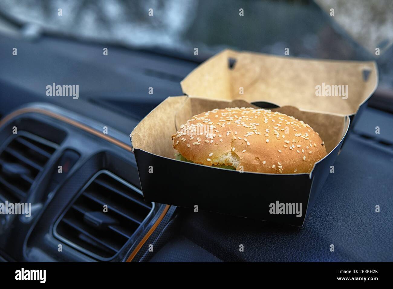Hamburger zum Mittagessen mit Bun und Sesam im Karton. Burger auf Konsole im Auto. Mittagspause bei Autofahrt, Mitnahme. Stockfoto