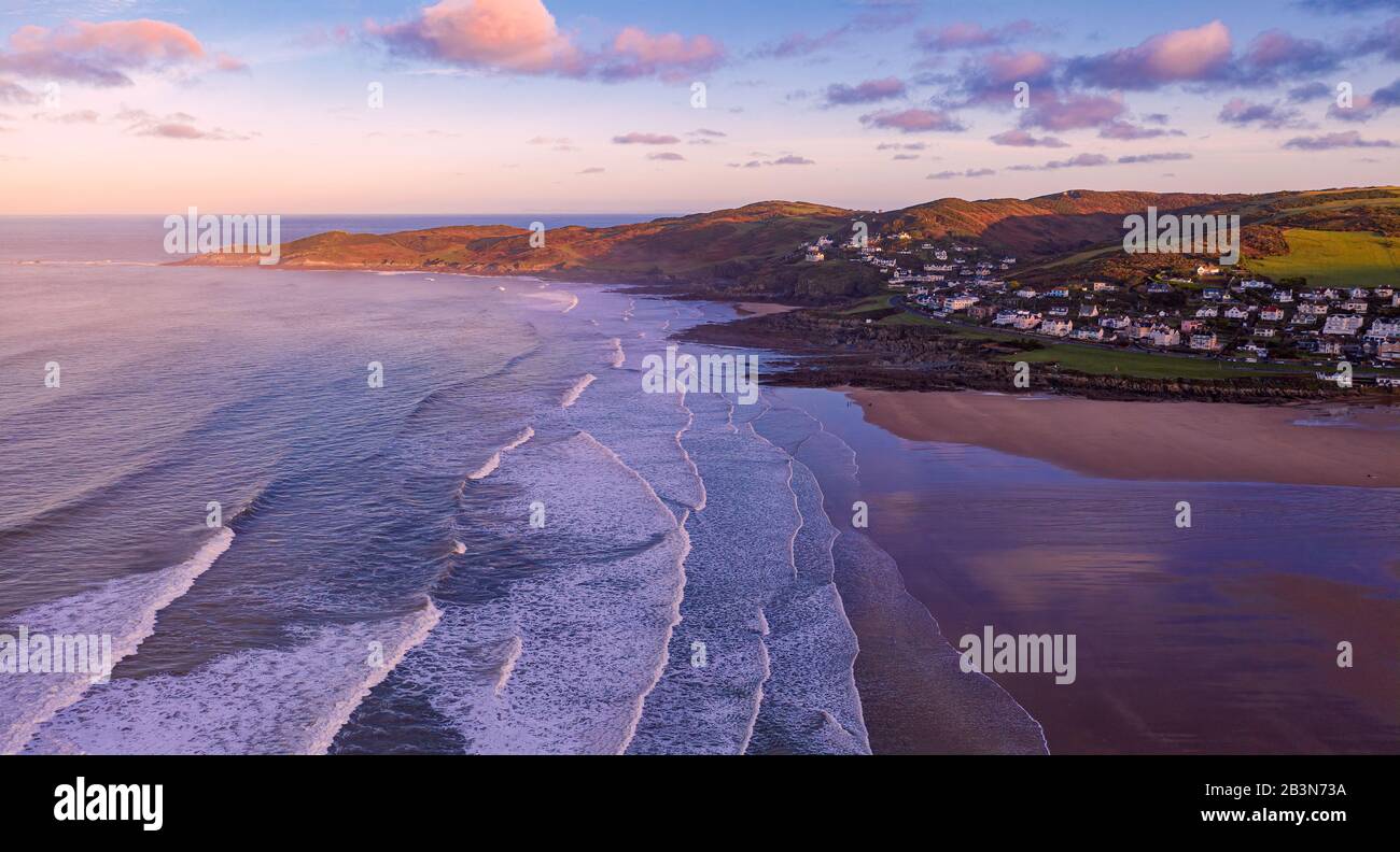 Luftaufnahme der Stadt Woolacombe und Strand im Morgengrauen, Wellen, die an einem Strand brechen, die Landzunge des Morte Point, die in Sonnenlicht und mit Hazy gebadet wird Stockfoto