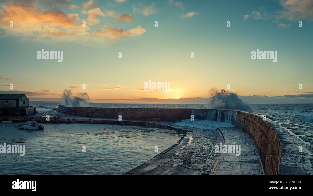 Wellen brechen über die Hafenmauer des cobb in Lyme Regis die Sonne, die gerade durch die Wolken am Horizont bricht und die Wolken malt Stockfoto