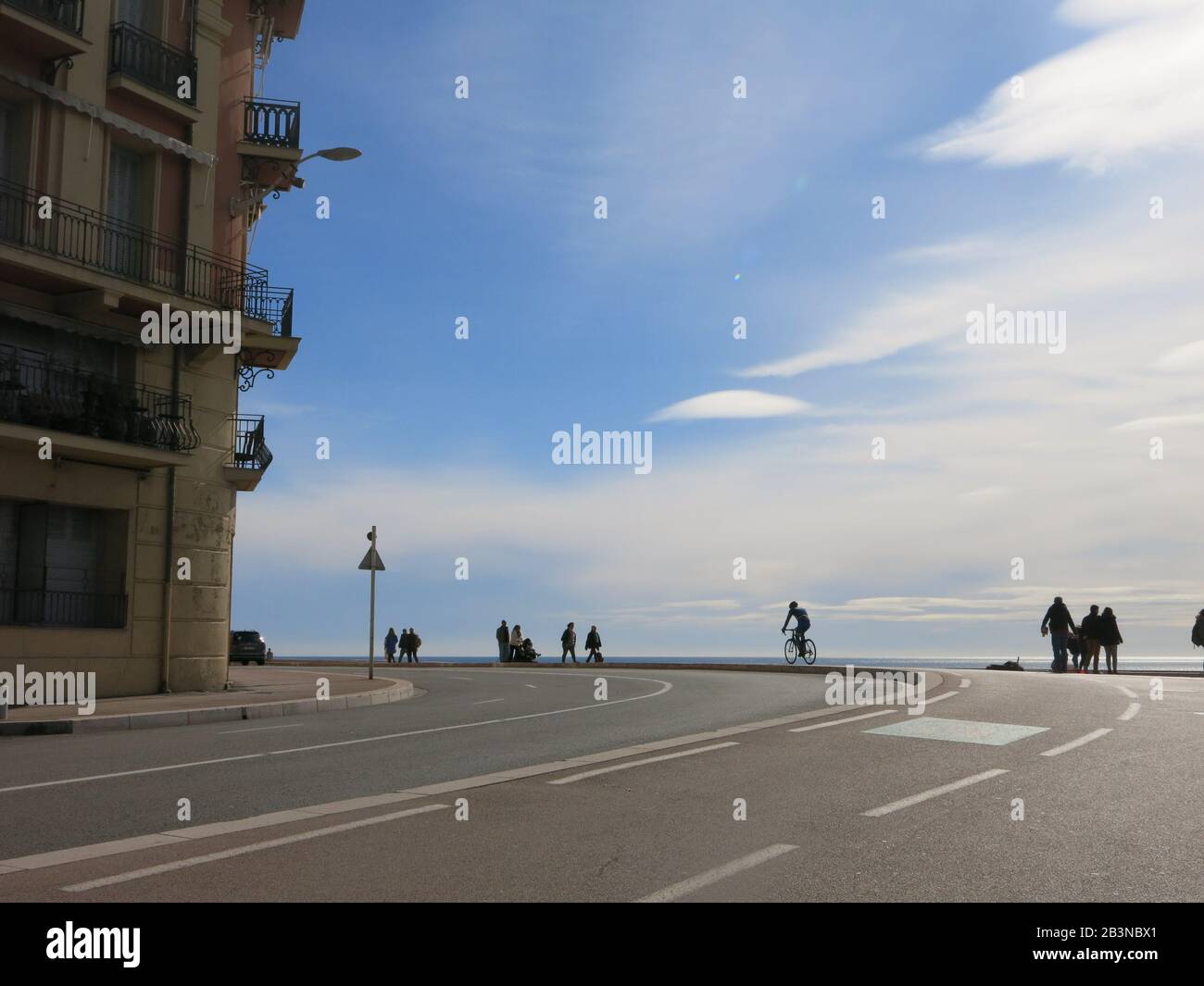 Die Straße biegt scharf nach links ab, während der Quai Rauba Capeu die Küste umarmt; Fußgänger und Radfahrer verschmelzen in den blauen Himmel des Horizonts. Stockfoto