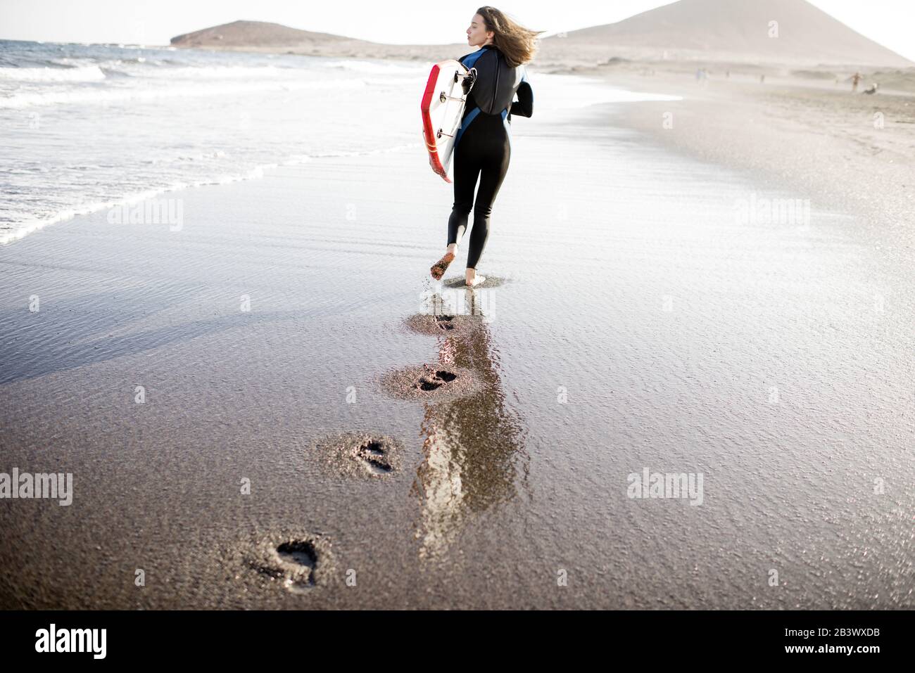 Junge Frau im Wetsuit, die mit Surfbrett spazieren geht, hinterlässt Fußabdrücke auf dem Sand, Blick von der Rückseite. Wassersport und aktives Lifestyle-Konzept Stockfoto