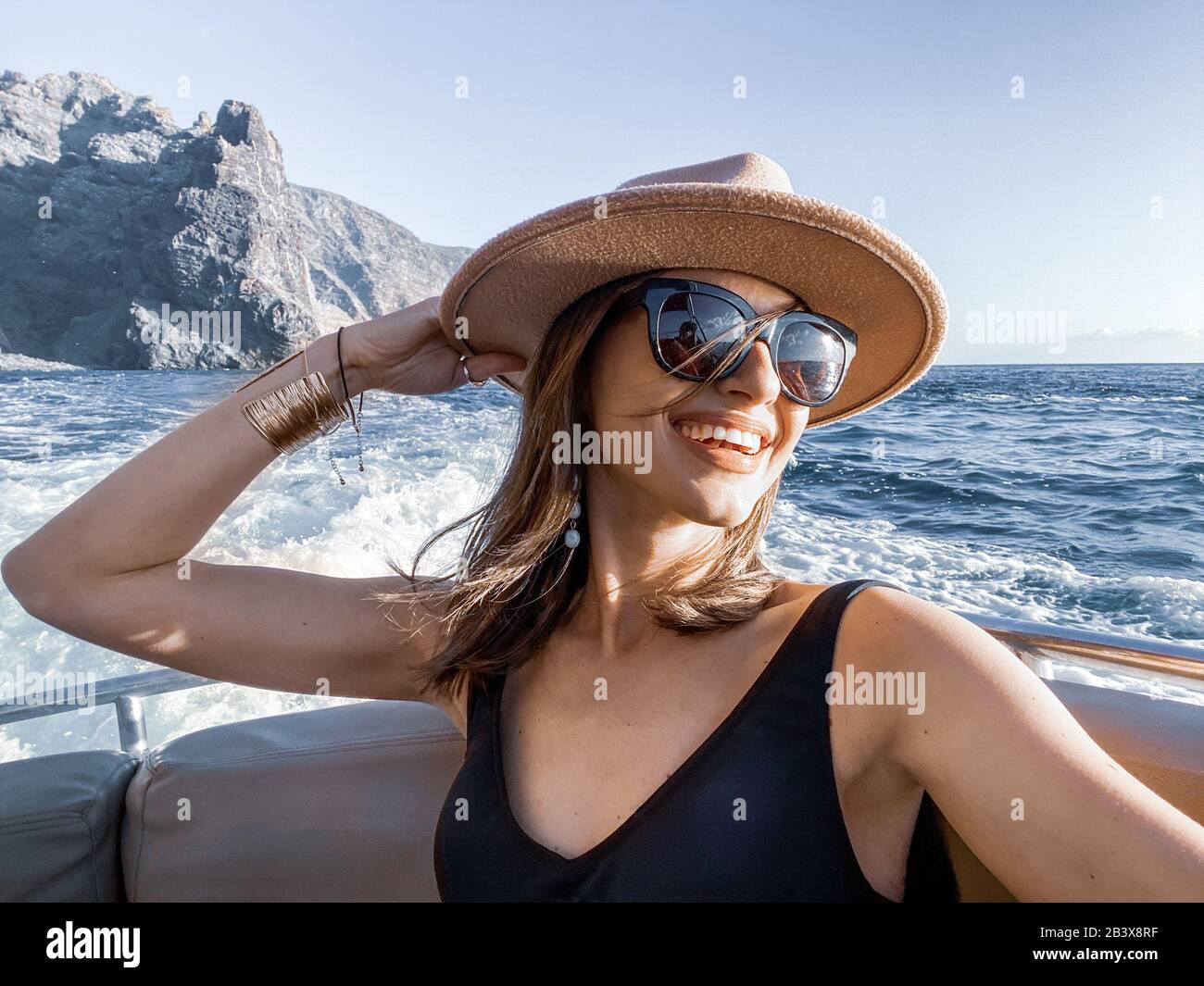 Frau im Badeanzug und Sonnenhut, die auf Ozeanfahrt geht, macht selfie auf dem Telefon mit schöner Felsküste im Hintergrund Stockfoto