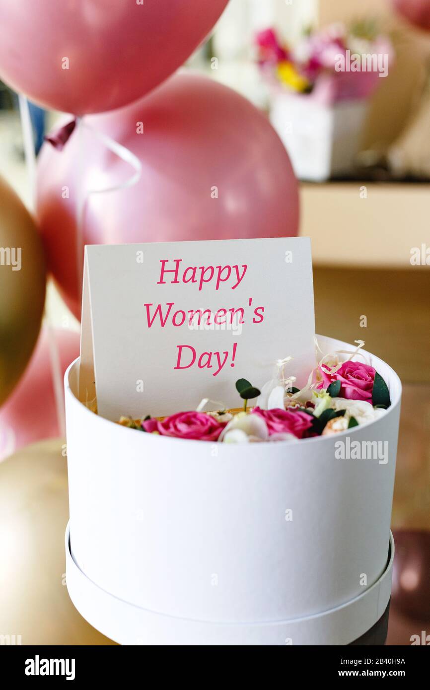 Happy Womens Day Text auf Geschenkkarte im Blumenkasten in der Nähe festlicher Rose und Goldballons. Grußkarte für Frauen am 8. März, internationaler Frauentag Stockfoto