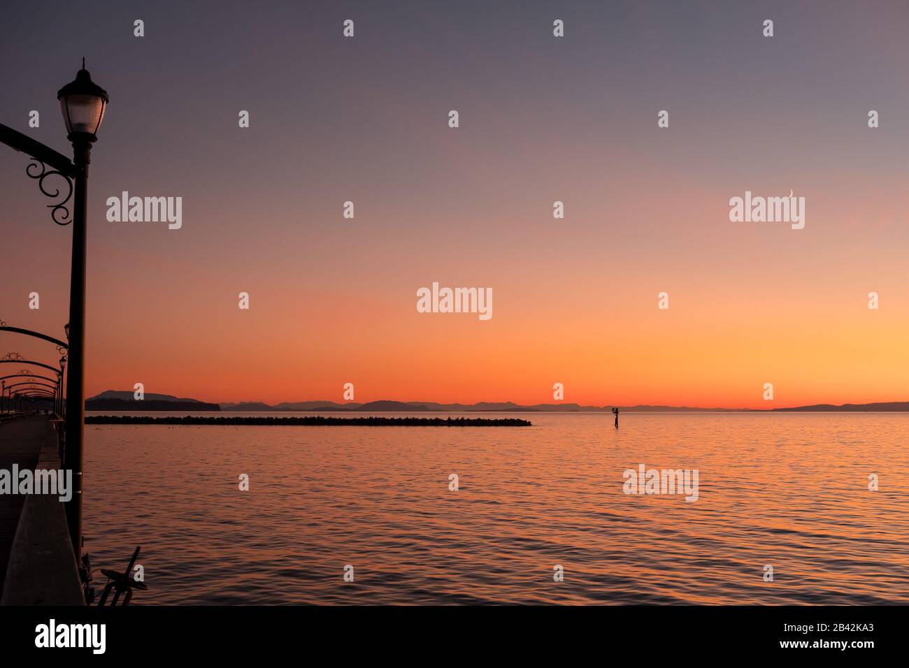 Der Himmel von Orangen, Rottönen, Pinks und Gelbtönen spiegelt sich in ruhigen Gewässern der Semiahmoo Bay, White Rock, BC, Kanada, wider. Oben schwebt ein Neumond in den Himmel. Stockfoto