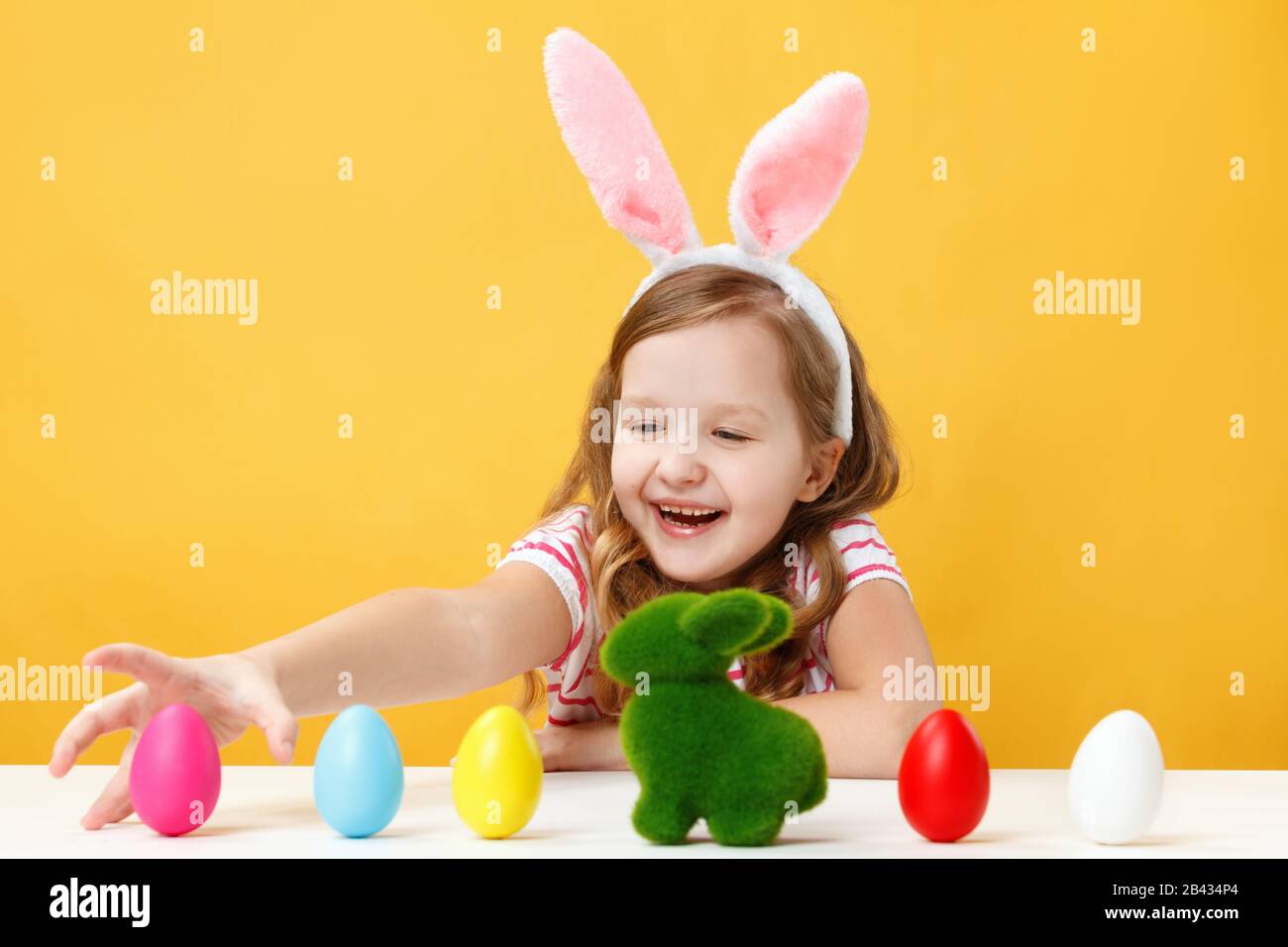 Osterkonzept. Glückliches Kind in den Hünenohren gelber Hintergrund. Ein kleines Mädchen sitzt an einem Tisch und jagt nach farbigen Eiern. Stockfoto