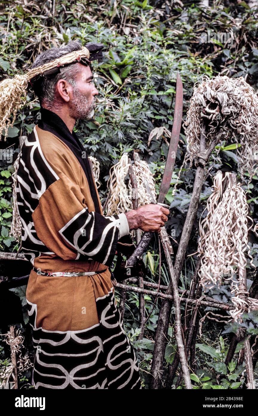 Ein bärtiger Ainu-Mann im traditionellen Kleid und mit einem Schwert führt eine Zeremonie auf einem Friedhof vor einem mit Holzspänen verzierten Schädel eines Bären auf der Insel Hokkaido im Norden Japans durch. Es lebten nur noch 300 reinblütige Ainu (ausgesprochen i-noo), als er 1962 für diese historische Fotografie posierte. Seitdem assimilieren sich die Ainu in die japanische Gesellschaft und ihre uralten Handwerke, Bräuche und Feierlichkeiten werden nur noch in besonderen Dörfern für Touristen am Leben gehalten. Früher als "haarige Aborigines" denigiert, wurden die Ainu 2008 offiziell als Ureinwohner Japans anerkannt. Stockfoto