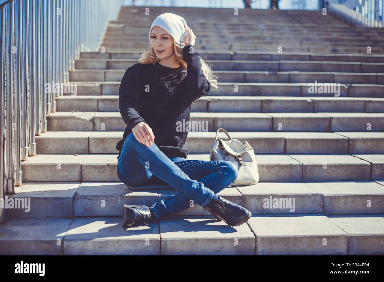 Modefoto einer jungen schönen Frau in Jeans, einem schwarzen Sweatshirt, einem weißen Strickmütze mit Rucksack in ihrem Showlder auf den Stufen eines s sitzend Stockfoto