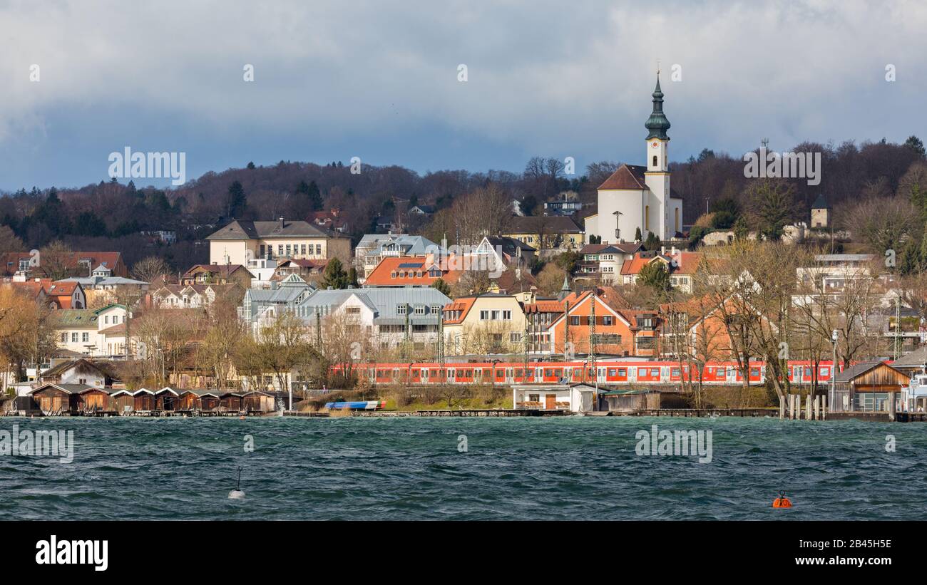 Blick auf die Stadt Starnberg. Mit der Kirche St. Josef (Sankt Josef), einem roten S-Bahn-Zug & Bootshaus. Im Vordergrund der Starnberger See. Stockfoto
