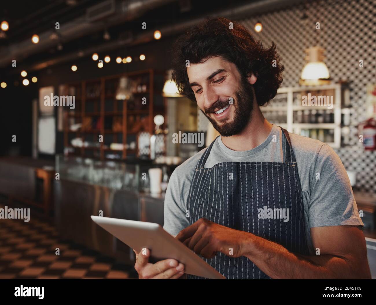 Fröhlicher junger kaukasischer Café-Besitzer, der Schürze mit digitalem Tablet trägt Stockfoto