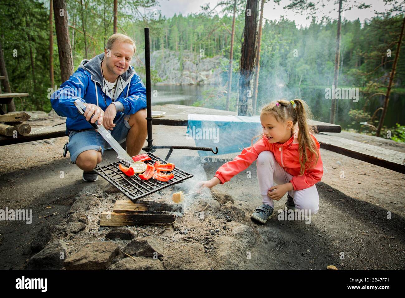 Der Mann und seine kleine Tochter grillen im Wald am felsigen Ufer des Sees und machen ein Feuer, grillen Brot, Gemüse und Marshmallow. Frohes Familienfest Stockfoto