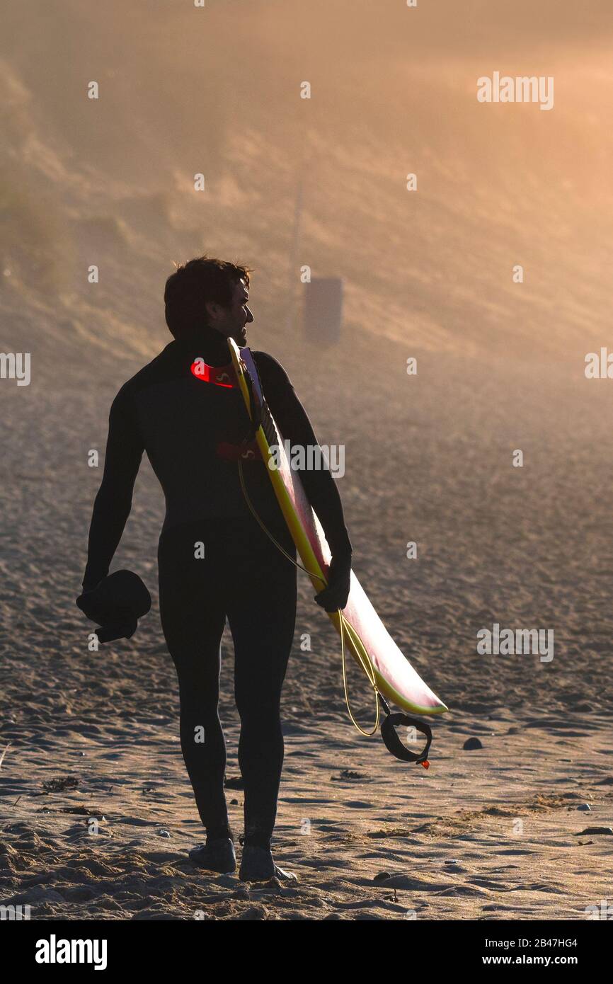 Ein Surfer, der sein Surfbrett trägt und am späten Abend in der Sonne am Fistral Beach in Newquay in Cornwall spazieren geht. Stockfoto
