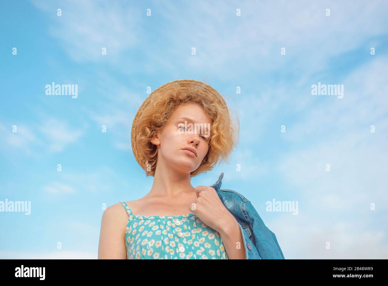 Junge, lockige Rotkopffrau in Strohhut, blaue Sundress und Jeansjacke auf blauem Himmelskleid. Spaß, Sommer, Mode, Schießen, Reisen, Jugend Stockfoto