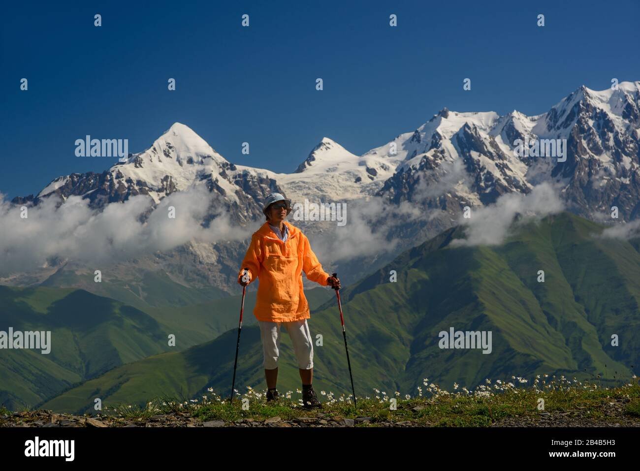 Tourist mit orangefarbener Jacke gegen Berge mit Schneehintergrund gekleidet. Alpenwiese mit weißen Blumen im Vordergrund. Stockfoto