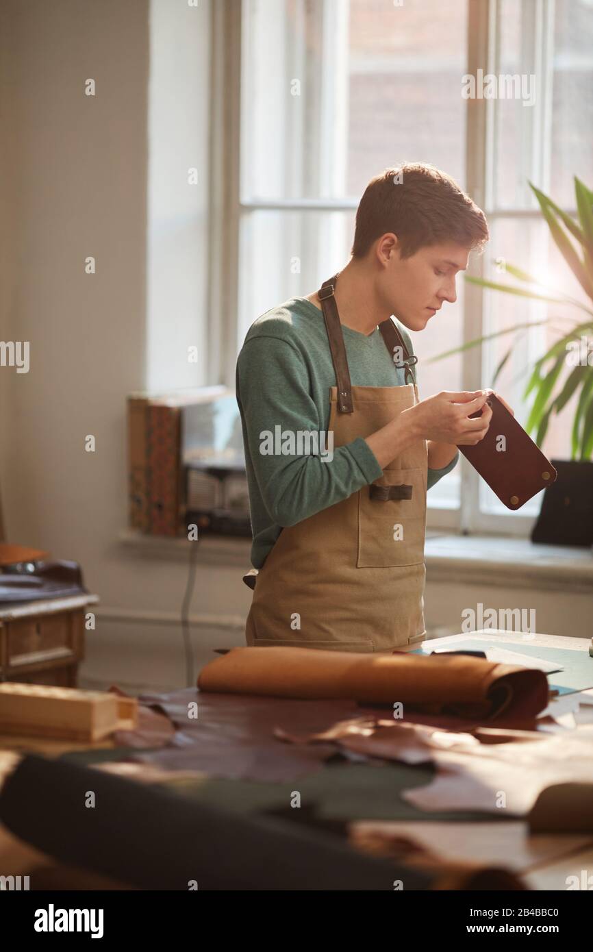 Vertikales, mittellanges Porträt junger Handwerker, die am Arbeitstisch stehende Schürze tragen und Lederdetails halten, um ihre Qualität zu überprüfen Stockfoto