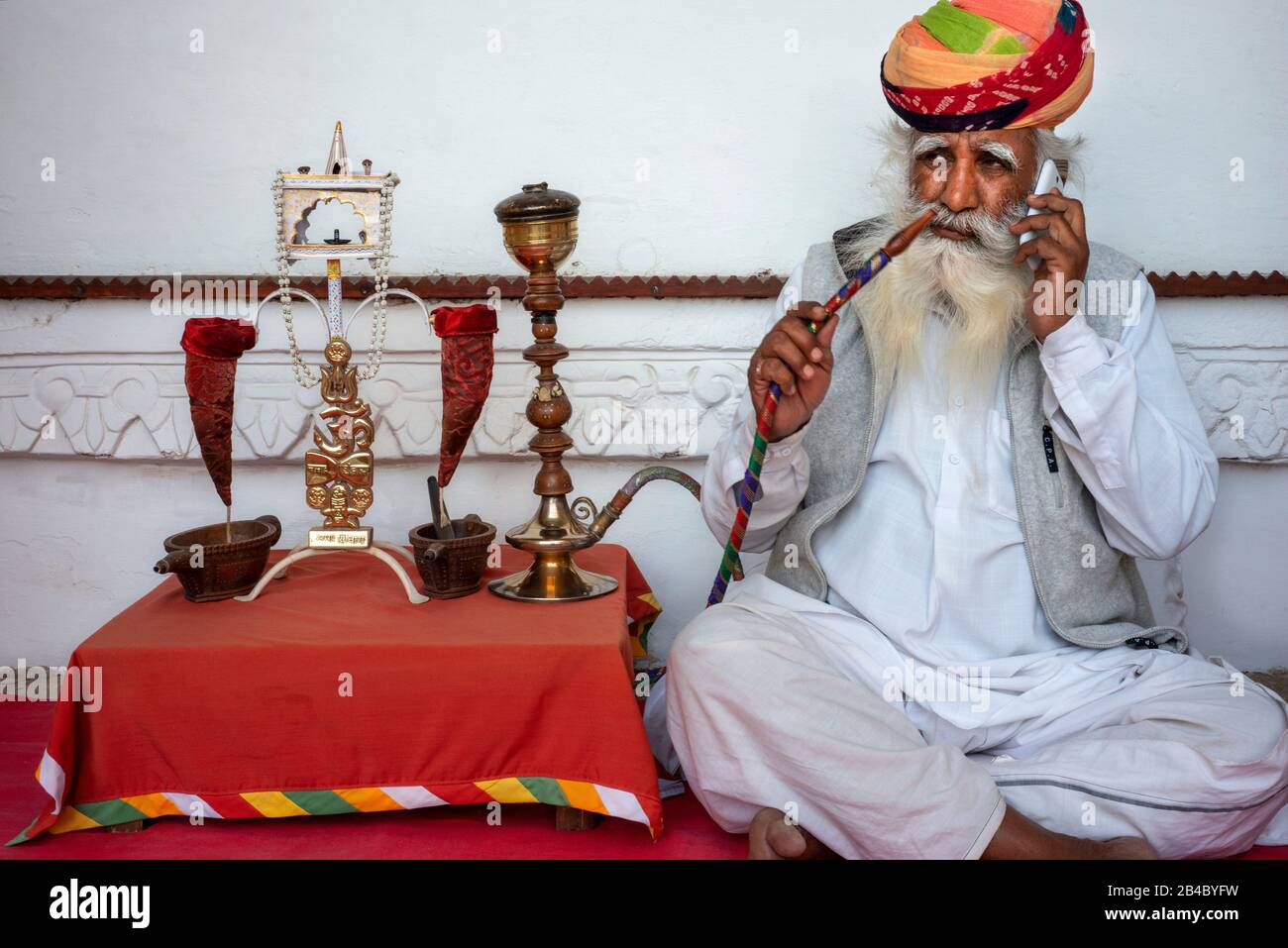 Gealterter indischer Mann mit langem weißem Bart und farbenfrohem Turban, der Opium aus einer Wasserleitung im Inneren von Mehrangarh Fort, Jodhpur, Rajasthan, Indien raucht. Das ist o Stockfoto