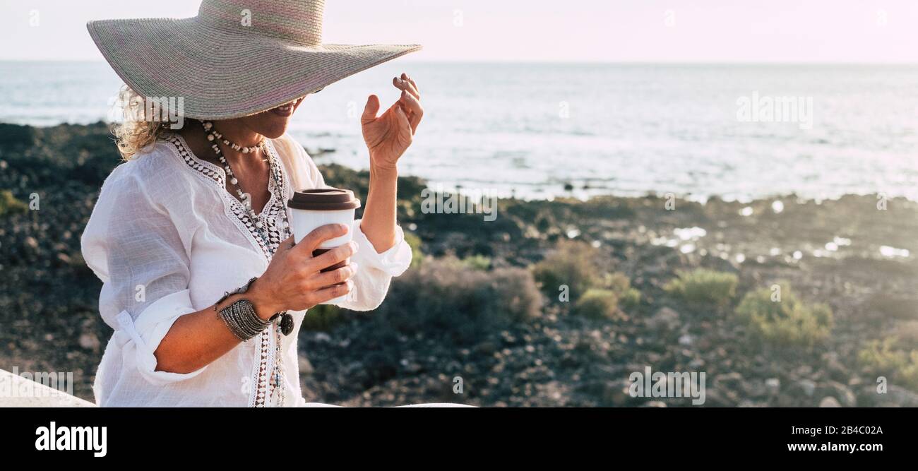 Modische trendige kaukasische Erwachsene Frau im mittleren Alter genießen die Freizeitaktivität im Freien beim Kaffeetrinken und Lächeln - Meer und Horizont im Hintergrund Stockfoto