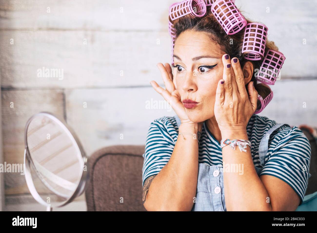 Schöne und lustige Ausdrucksweise kaukasische Erwachsene Frau, die sich zuhause vor dem Spiegel mit Schminke auf dem Gesicht - Falten prüfen und älteres Konzept für junge Leute bekommen - Lockenstab auf dem Haar Stockfoto