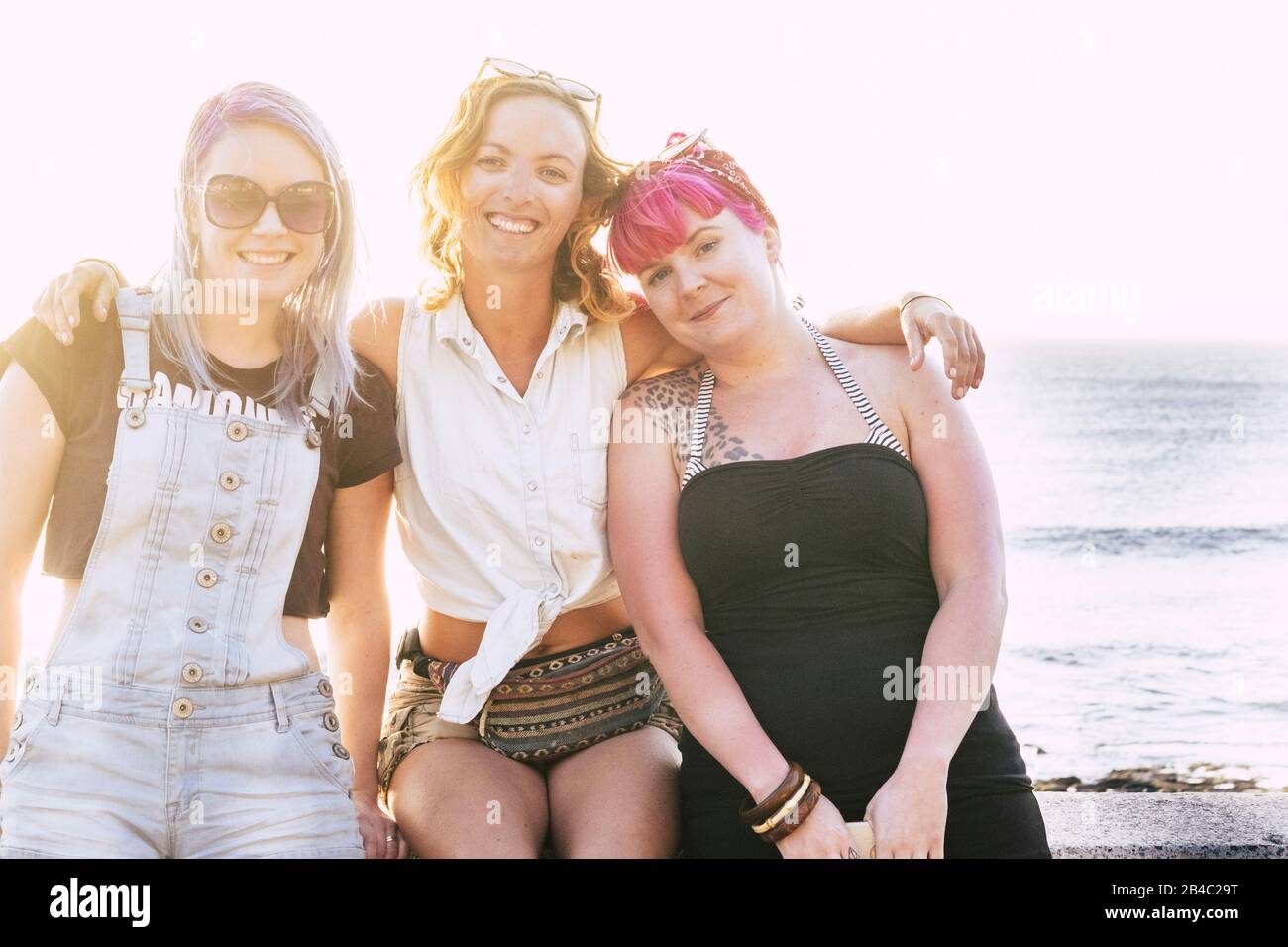 Drei Frauen umarmten und sitteten am Strand, schauten und lächelten mit der Kamera am Meer und dem blauen Wasser im Hintergrund - Freundschaftszielkonzept Stockfoto