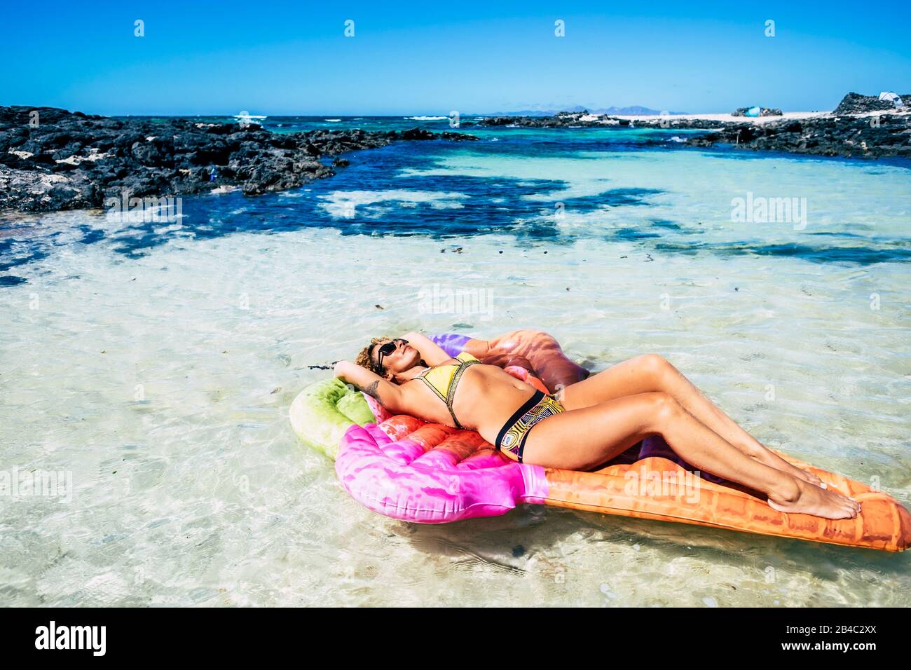 Schöne junge Frau hat ein Sonnenbad, das sich auf einer trendigen lilo aufblasbaren Matratze in einer blauen tropischen Ozeanlagune mit Sand und Felsen entspannen kann - Hafen- und Paradieskonzept für Reisen und Lifestyle Stockfoto