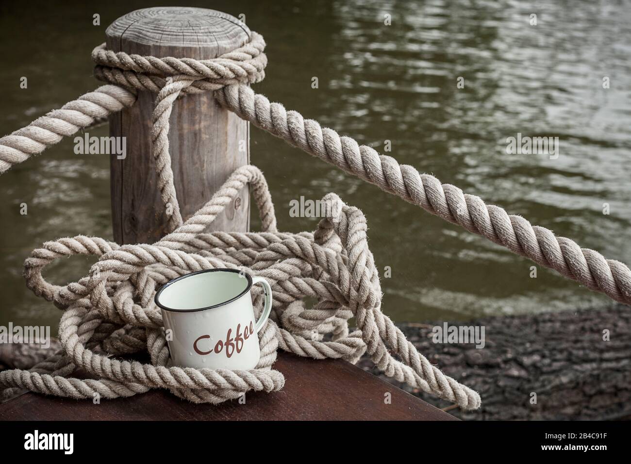 Rustikale Tasse aus Email mit Wortkaffee und dekorativem dicken Seil an einem Holzsteg am Wasserrand Stockfoto