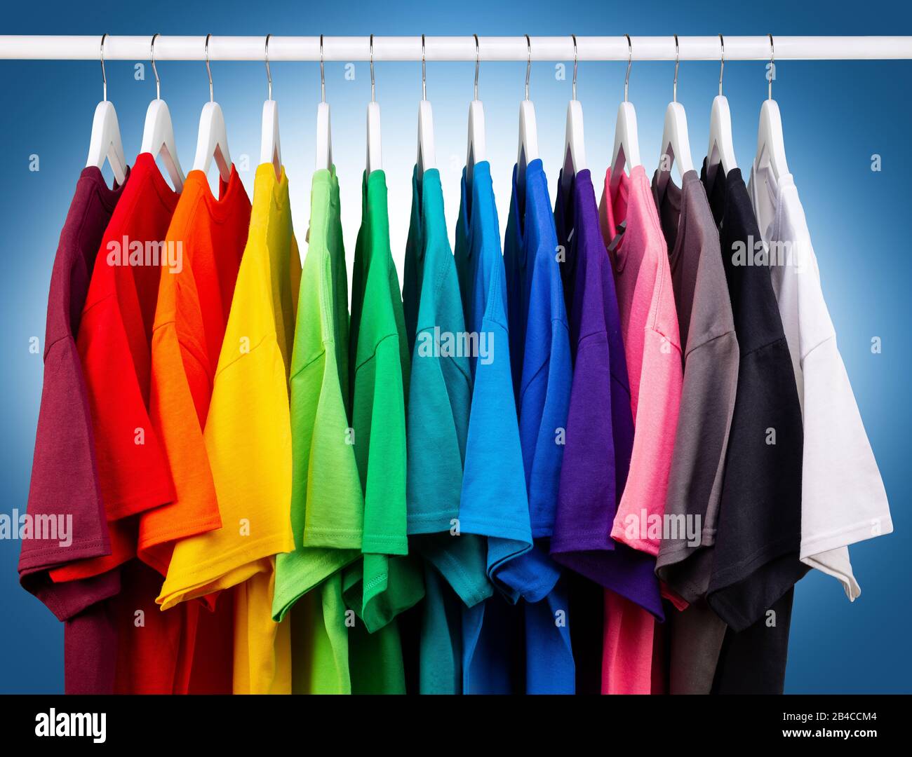 Viele neue, frische T-Shirts aus Baumwolle in bunten Regenbogenfarben hängen auf der Kleiderstange im Wrack. Verschiedene farbige Hemden auf blauweißem Backgr Stockfoto