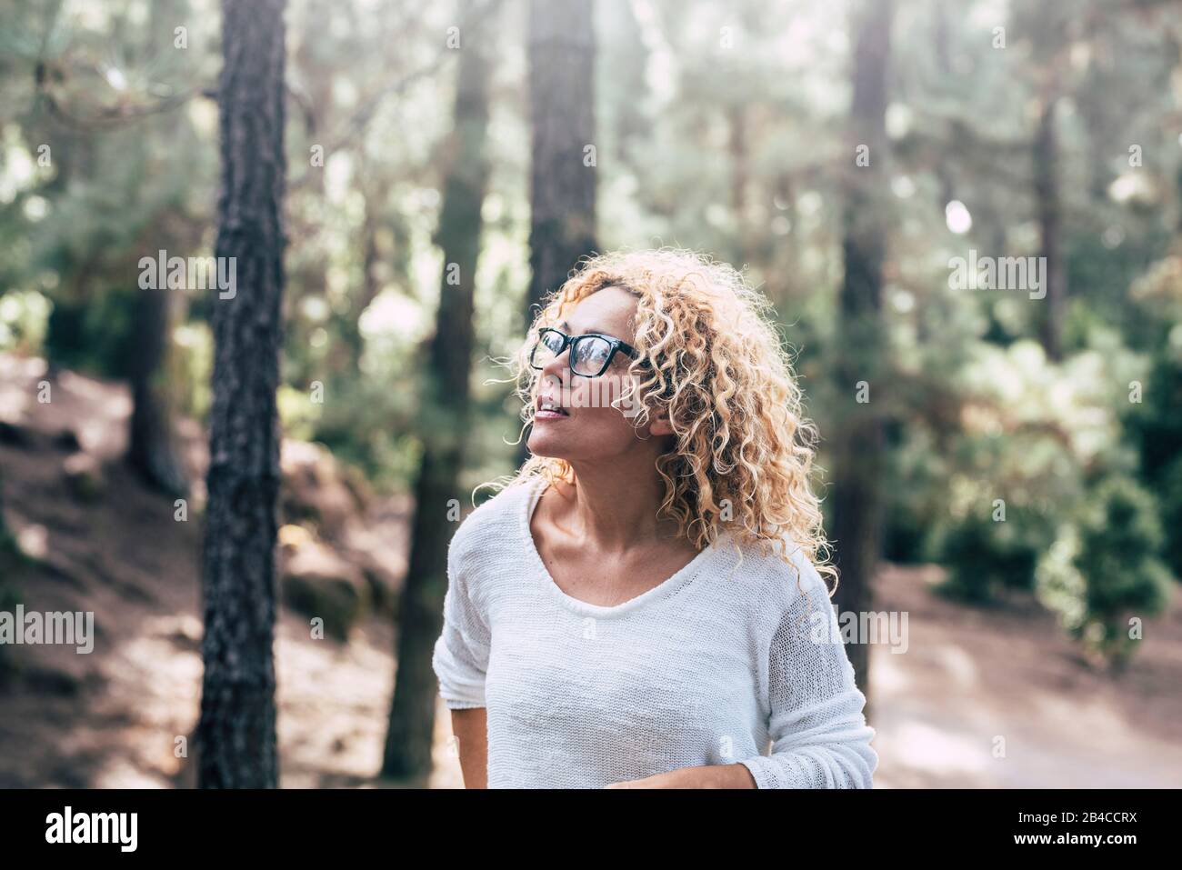 Schöne blonde Curly Woman genießen die Freizeitgestaltung im Wald im Freien - Menschen in den Holzbäumen Natur mit unschärfem Hintergrund und Sonne in Hintergrundbeleuchtung Stockfoto
