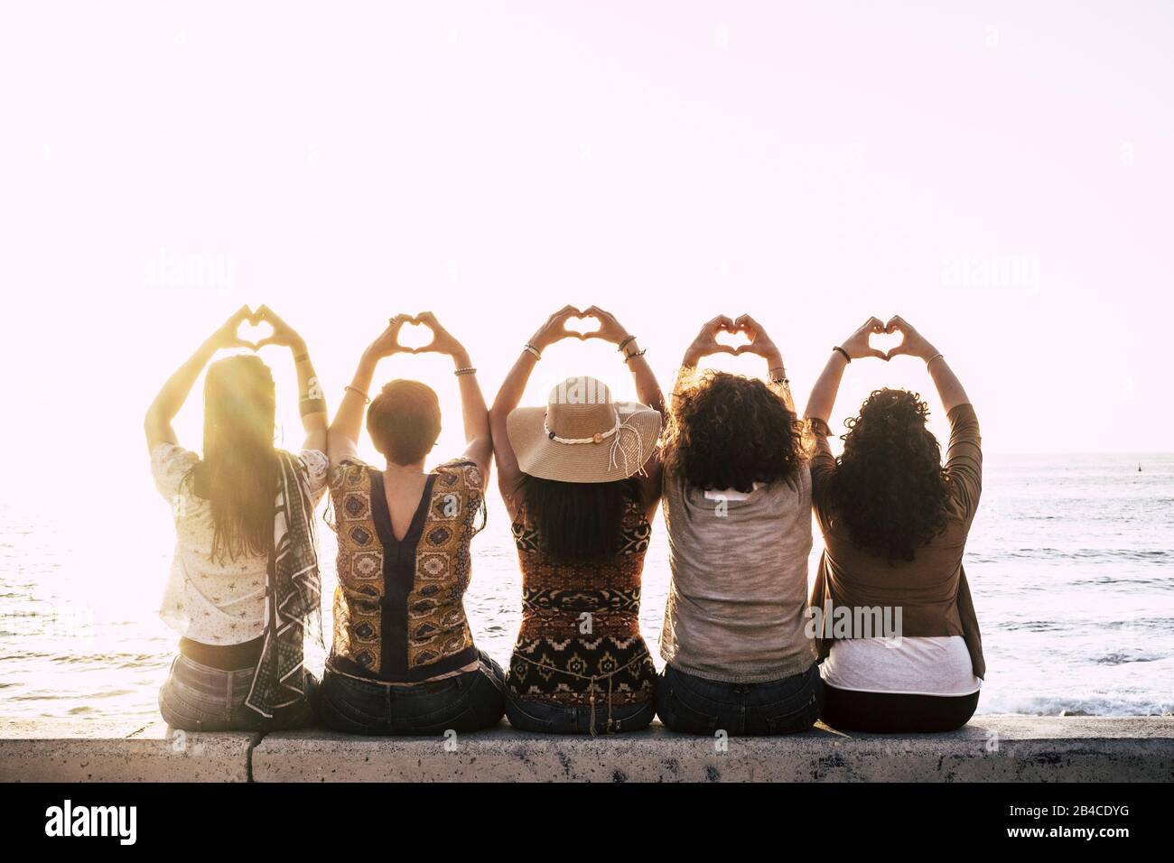 Eine Gruppe glücklicher Menschen im Freien, die Frauen von hinten gesehen haben, die ein Zeichen des Liebeskummer mit den Händen nach oben machen - Beziehungskonzept - Sonnenuntergang und Meer im Hintergrund Stockfoto