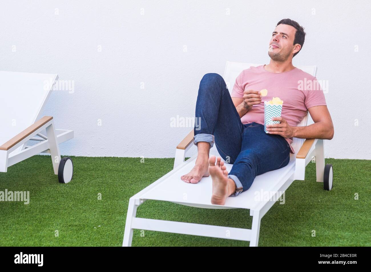 Gutaussehender einfacher Mann, der zu Hause Trödel fod isst, legte sich in der Sonnenliege nieder - genießen Sie die Entspannungsaktivität mit künstlichem Wiesengras und weißem minimalem Backgorund Stockfoto