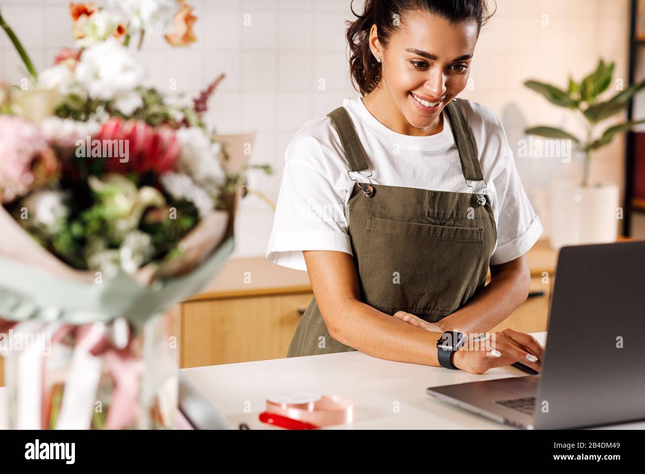 Fröhlicher Blumenhändler, der an der Theke arbeitet. Weibliche Floristin prüft Online-Bestellungen auf einem Laptop. Stockfoto