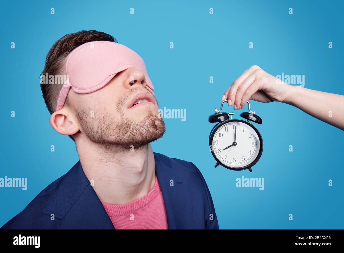 Weibliche Hand hält den Wecker mit Schlafmaske an den Augen nahe am jungen Mann, während er vor blauem Hintergrund nappt Stockfoto
