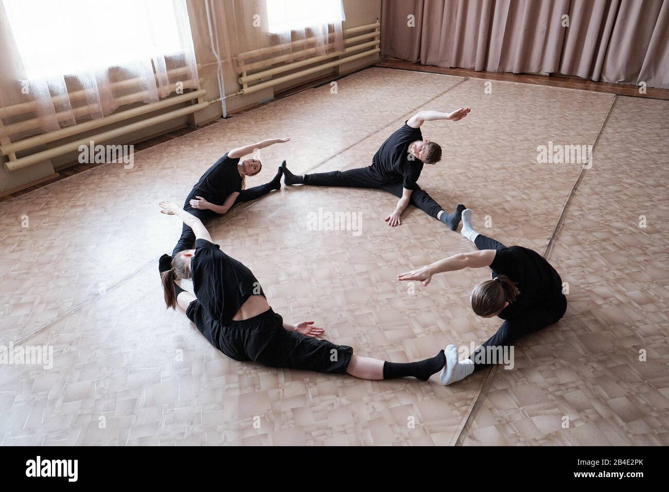 Vier junge, flexible Tänzerinnen in Avtivewear sitzen mit ausgestreckten Beinen während des Trainings auf dem Boden des modernen Tanzstudios Stockfoto