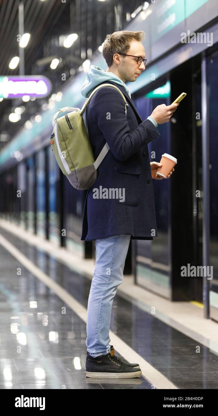 Kaukasischer junger Mann mit Rucksack mit Handy, der am frühen Morgen auf den Zug am Bahnsteig der U-Bahn wartet, eine Tasse Kaffee hält, geht zu w Stockfoto