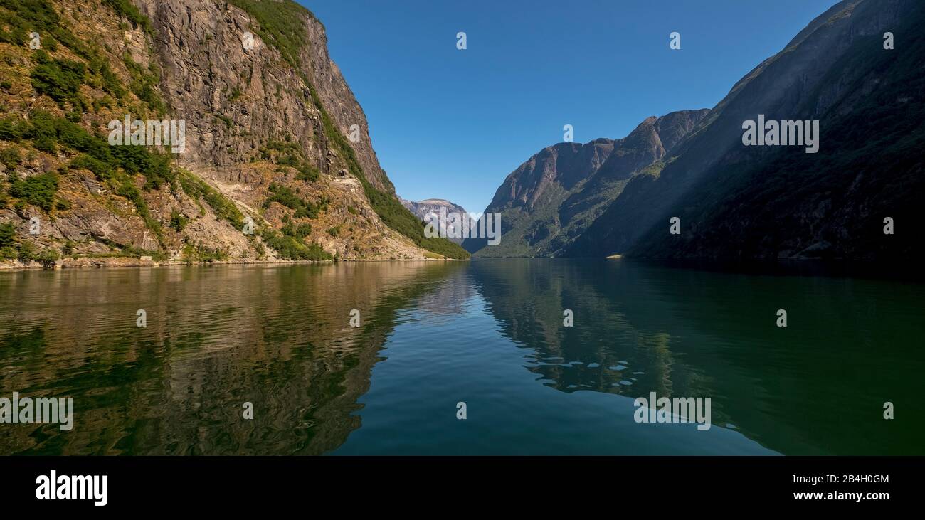 Ein schöner Fjord, in dem das Licht reflektiert wird, umgeben von Felsen, die teilweise mit Bäumen bewachsen sind, darüber ein strahlend blauer Himmel. Gudvangen, Sogn og Fjordane, Norwegen, Skandinavien, Europa Stockfoto