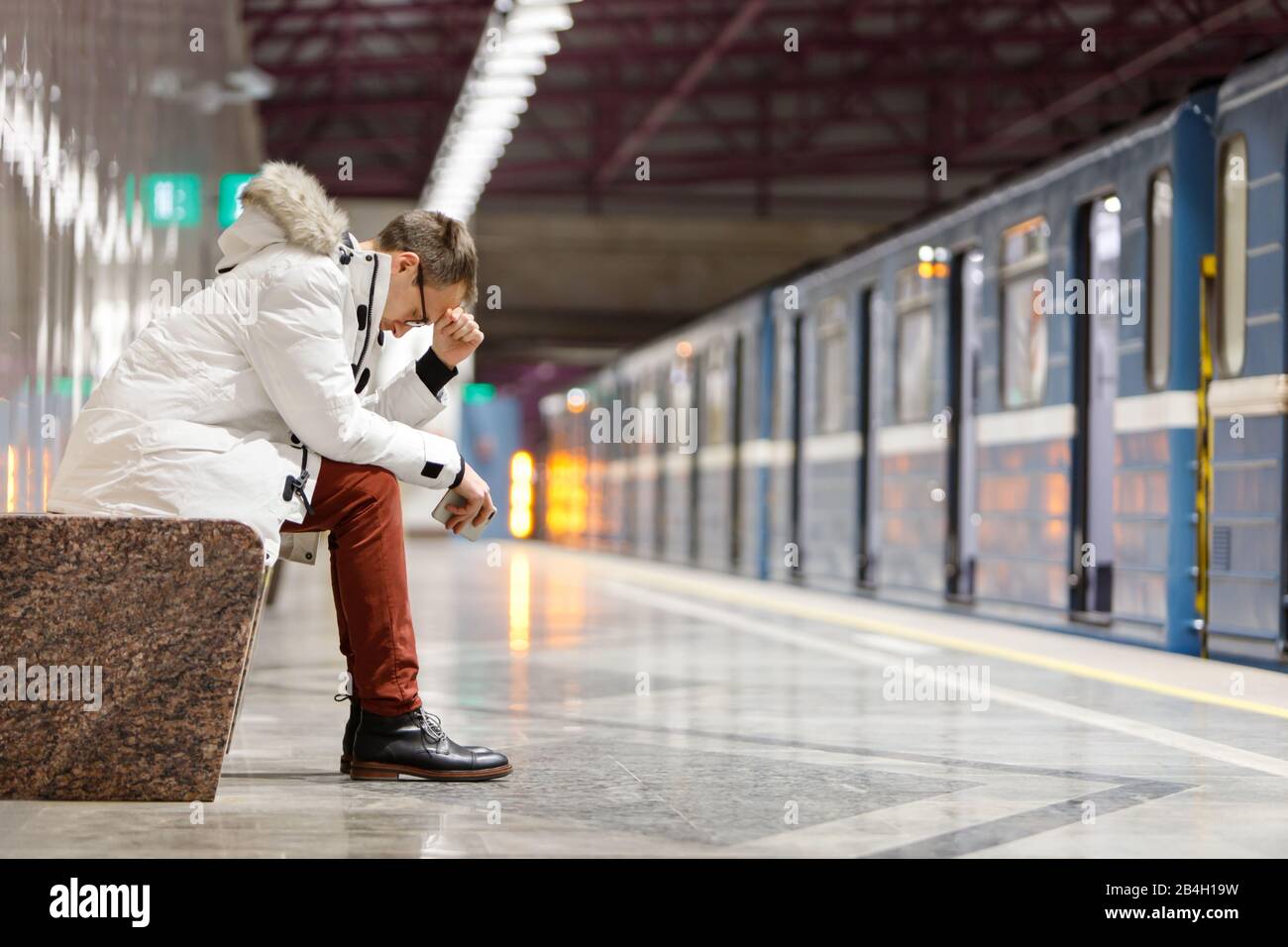 Weinender Mann in weißer Parka sehr verärgert, hält Smartphone, bekommt schlechte Nachrichten, hält mit der Hand die Stirn und sitzt auf der Bank in der U-Bahn-Station. Problem in Stockfoto