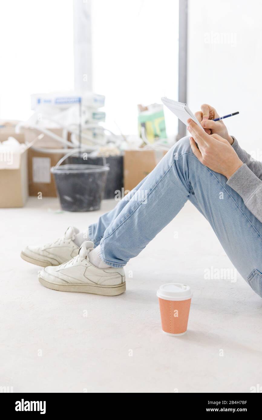 Der Mann hat eine Kaffeepause von der Renovierung, sitzt auf dem Boden und schreibt anstehende Einkäufe in seinem Notizbuch, ruht nach der Arbeit, weiche Fokussierung. Home RE Stockfoto