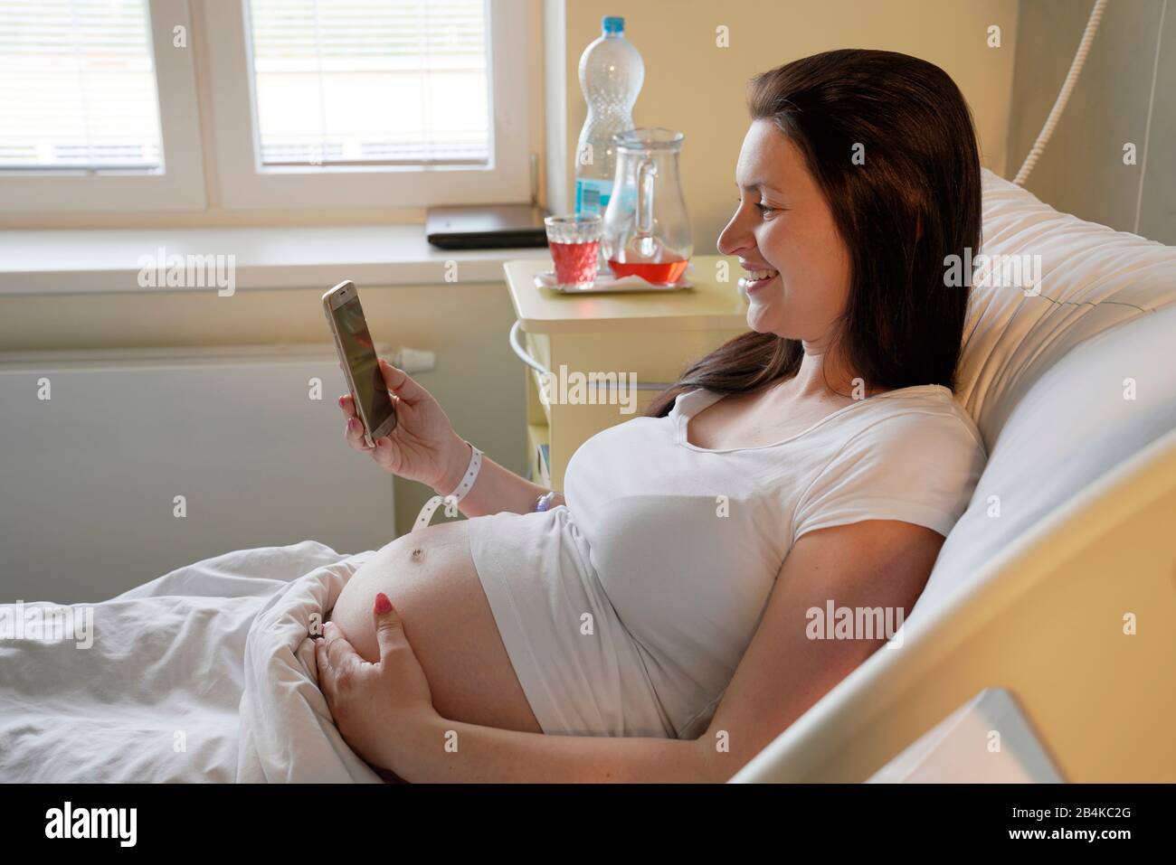 Hochriskige Schwangerschaft, schwangere Frau, die im Krankenhaus auf dem Mutterschaftsbett liegt und auf ihrem Handy sucht Stockfoto