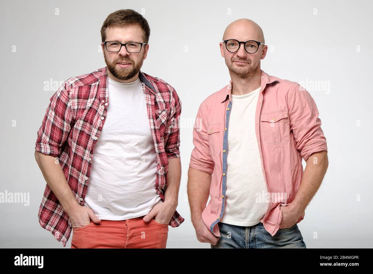 Zwei gutaussehende Männer in einer Brille, die ihre Hände in den Taschen halten und ruhig auf die Kamera blicken. Stockfoto