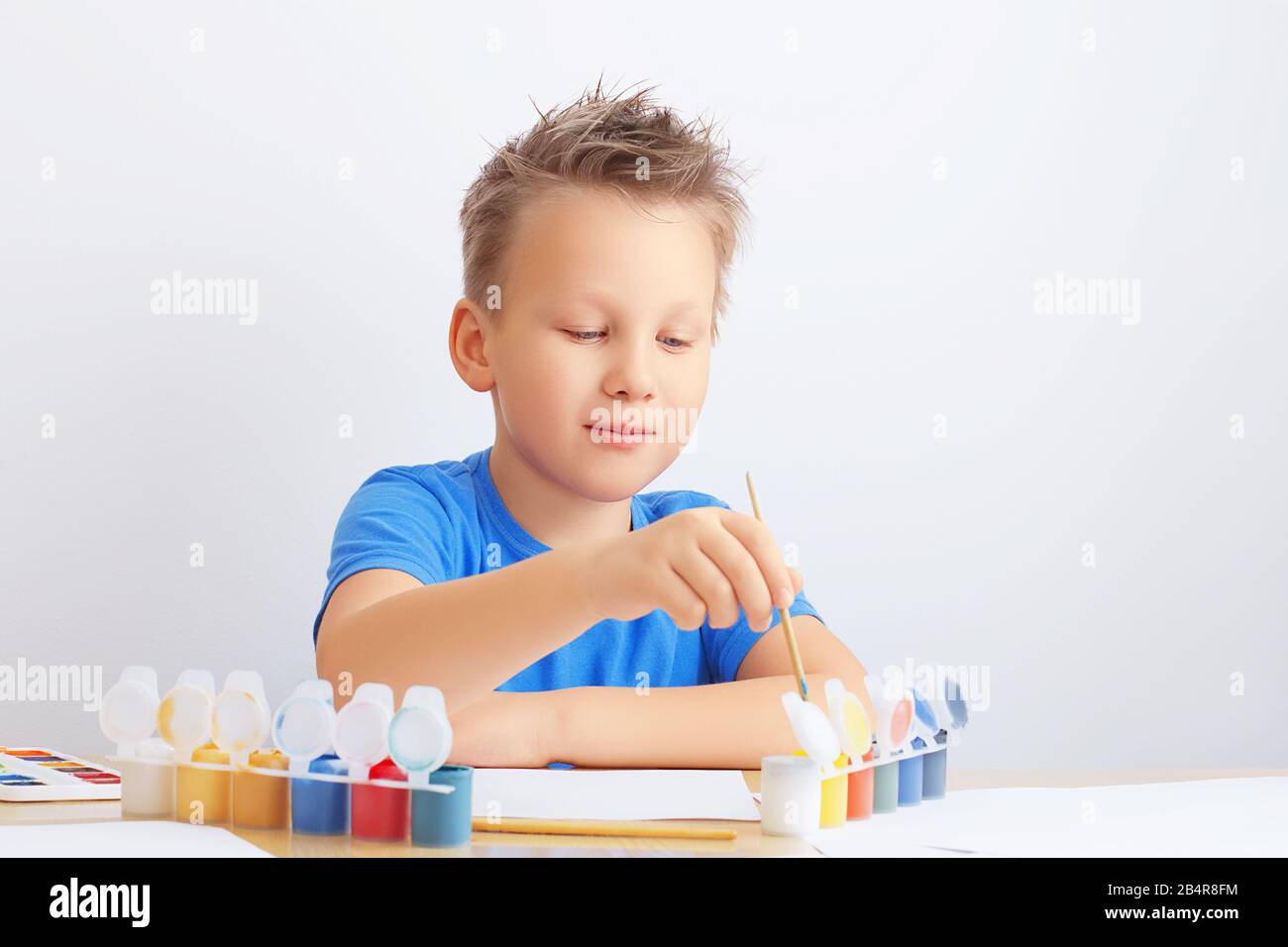 Ein Junge mit unordentlichen Haaren taucht einen Pinsel zum Malen in eine Dose Farbe. Schule, Kindergarten Kunstbildungskonzept Stockfoto