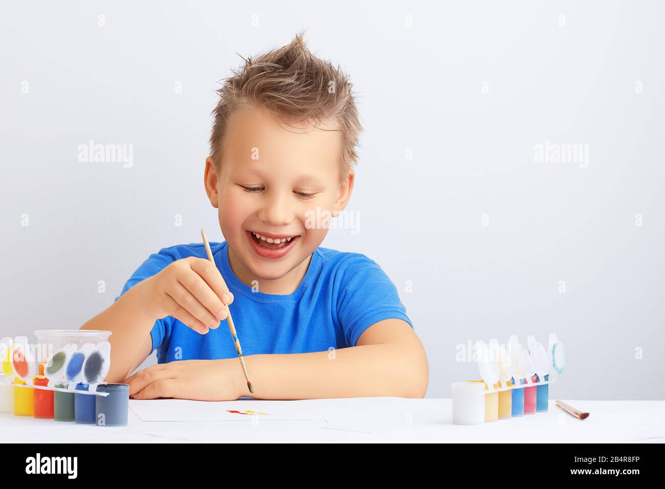 Ein fröhlicher lachender kleiner Junge mit unordentlichen Haaren hält einen Malpinsel in der Hand und malt mit bunten Farben auf einem weißen Blatt Papier. Schuleduc Stockfoto
