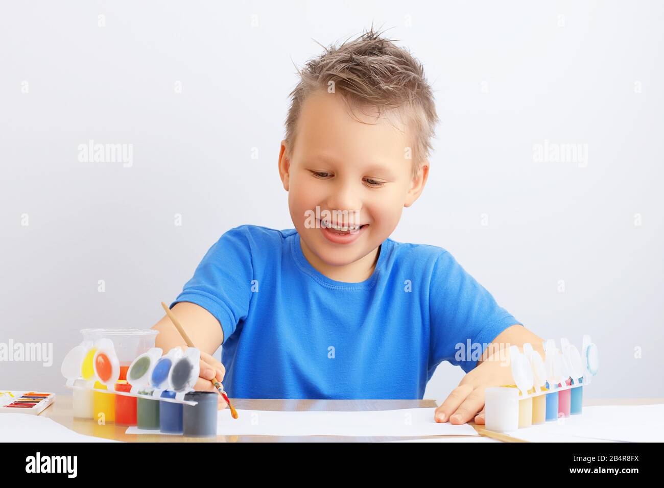 Ein fröhlicher lächelnder Junge mit unordentlichen Haaren hält einen Pinsel in der Hand und zieht mit farbigen Farben auf Papier. Kreativität, Bildung, Kunstkonzept. Stockfoto